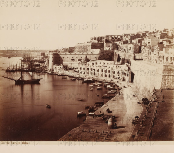 View of the harbor of Malta, Baracca-Malta, photograph, inscription: '(+) Sommer', number: 'No. 1 12.', photographer: attributed to Giorgio Sommer, Malta, 1857 - 1914, Attributed to, pasteboard, paper, albumen print, height 270 mm x width 355 mm, harbour, city-view in general, 'veduta', quay, A wide waterfront scene shows a calm harbor with several sailing ships at anchor and smaller rowboats along the quay, their masts and rigging reflected in the water; a fortified stone embankment rises from the water, topped by terraces and a densely packed cluster of multi-story buildings with balconies and arched openings; a promenade along the quay features scattered barrels, carts, and small bundled goods, with a series of stone posts and decorative balustrades lining the upper walkway; architectural details include large rounded bastions, arched openings at water level, and a prominent façade with ornate stonework set into the fort wall; distant low hills and open shoreline are visible across ...