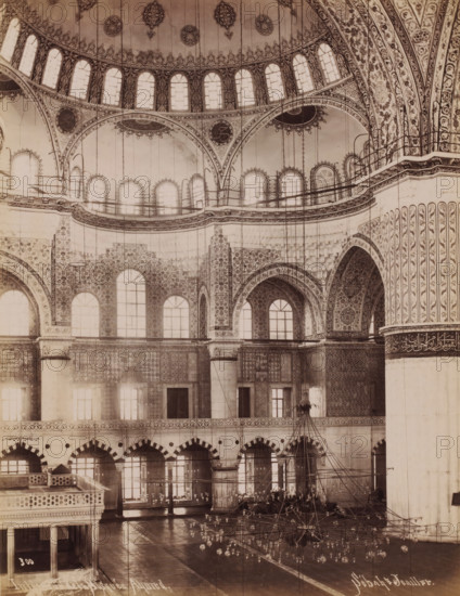 Interior of the Sultan Ahmet Mosque in Istanbul, Intérieur de la Mosquée Ahmed, photograph, number: '300', photographer: Sébah & Joaillier, Istanbul, 1888 - 1900, paper, albumen print, height 268 mm x width 208 mm, temple, shrine, Islam, Mohammedanism, Sultan Ahmet Mosque, The image shows an interior space with a large ornate dome and multiple tiers of arched windows and balconies. Intricate floral and geometric patterns cover the walls, arches, and columns, with decorative medallions and ornate borders visible along curved surfaces. Tall slender columns rise to support arches adorned with detailed motifs, and a series of hanging chains and light fixtures descend from the ceiling toward a central chandelier arrangement near the floor. The lower wall surfaces display patterned tilework and rows of arched doorways or niches, while the floor appears polished and reflective. A partial elevated wooden structure with balustrades is visible in the foreground, and a large plain structural pill...