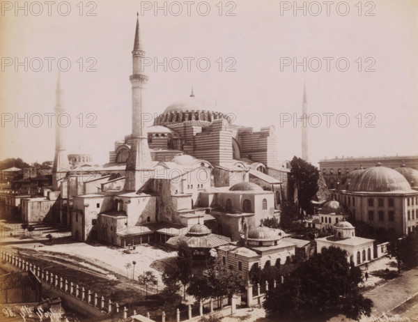 View of the Hagia Sophia in Istanbul, Ste Sophie, photograph, number: '98.', photographer: Sébah & Joaillier, Istanbul, 1888 - 1900, paper, albumen print, albumendruk, height 208 mm x width 268 mm, temple, shrine, Islam, Mohammedanism, Hagia Sophia, A large domed building with striped exterior walls fills the center of the image, surrounded by multiple smaller domes, vaulted roofs, and rectangular structures clustered tightly around it; tall slender minarets rise from the complex, one prominent in the foreground and others visible toward the edges, each with a pointed top and a narrow shaft. The foreground shows a paved open area with scattered trees and a low decorative fence running along the bottom, while additional domed buildings and an arched colonnaded structure appear to the right. Rooflines, staircases, and small annexes attach to the main building, and a few trees and patches of vegetation are interspersed among the stone and plaster structures. The scene is rendered in a pal...