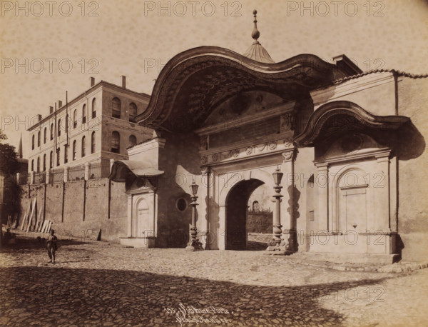 Sublime Porte in Istanbul, Sublime Porte, photograph, number: '195.', photographer: Sébah & Joaillier, Istanbul, 1888 - 1900, paper, albumen print, albumendruk, height 208 mm x width 268 mm, gate, entrance, A sepia-toned street scene showing a large ornate arched gateway with carved decoration and a curved, painted canopy above, flanked by smaller doorways and ornate lamp posts on stone pedestals; a multi-story building with arched windows and a fenced parapet sits behind a high wall to the left, with wooden planks leaning against the wall and a narrow spire visible in the distance; a cobblestone street fills the foreground where a solitary child stands wearing simple clothing and holding an object, casting a shadow on the ground.