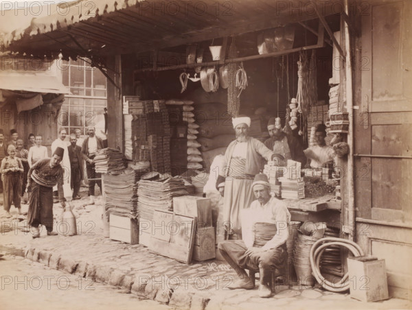 Brousse Marchands kurds, View of a Kurdish shop in Bursa, photograph, number: 'No, 111.', photographer: Sébah & Joaillier, Bursa, 1888 - 1900, paper, albumen print, albumendruk, height 205 mm x width 269 mm, shop, store, A crowded market stall under a corrugated awning shows stacks of folded textiles and piled boxes on wooden crates in front of a dark interior filled with sacks and hanging bundles. Several men in traditional garments and headwear stand and sit among the goods, one seated on a small stool near coils of hose and metal buckets, another standing with a hand resting on stacked boxes, and others arranged toward the left and background near a windowed building. Metal pans, ropes, and long strands of dried items hang from a horizontal beam above the stall, and a man in the foreground bends slightly while holding a large metal container on the cobbled street. The scene includes wooden doors and shelving, assorted small packaged items on display, and a stone curb bordering the u...