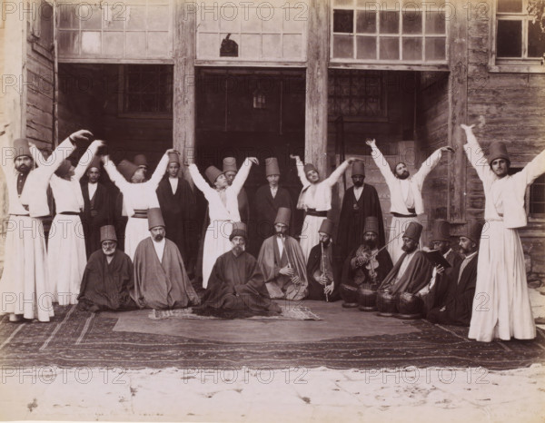 Dervishes during a sama, photograph, inscription: 'illegible', photographer: Sébah & Joaillier, Middle East, 1888 - 1900, paper, albumen print, height 210 mm x width 270 mm, group dancing, A group of people arranged in front of a wooden building with large paned windows, some standing with arms raised and some seated on carpets on the ground; many wear tall cylindrical hats and loose robes, a few hold musical instruments including wind instruments and a bowed instrument, and several seated figures have long outer garments while others in the back wear lighter robes and belts, with visible wooden pillars and window frames behind them and patterned rugs beneath their feet.