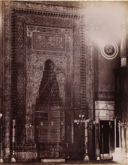 Interior of a mosque, possibly in Istanbul, photograph, photographer: attributed to Sébah & Joaillier, Istanbul, 1888 - 1900, Attributed to, paper, albumen print, albumendruk, height 270 mm x width 210 mm, temple, shrine, Islam, Mohammedanism, A sepia-toned photograph showing a section of a richly decorated interior wall with an ornate recessed niche framed by intricate geometric and floral patterns, scalloped arch detailing, and layered rectangular panels. The wall surface is covered in dense repeated motifs and borders, with vertical columns on either side that have carved or painted decorative bands. A large round medallion with concentric rings and an emblematic mark is visible on the right above a plain wall area. Hanging lamps and chandeliers suspended by thin wires or cords appear across the foreground, and tall slender columns with bases and capitals stand near a recessed doorway on the right. The lower portion shows a raised platform or step and detailed friezes along the base...