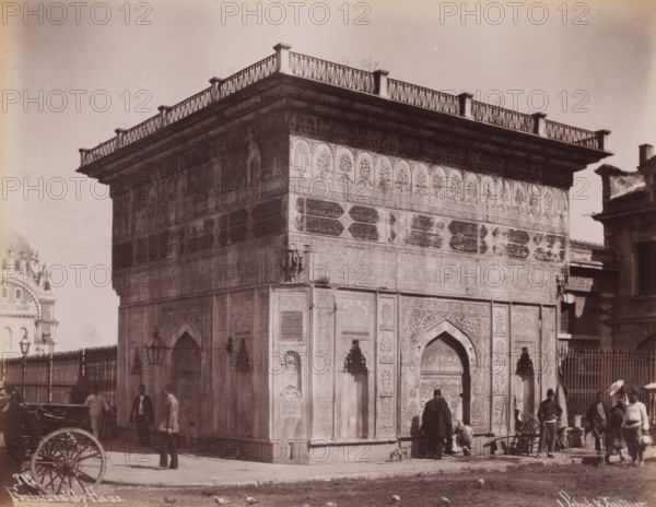 Fontaine a Top-Hane, Tophane fountain in Istanbul, photograph, number: '171.', photographer: Sébah & Joaillier, Istanbul, 1888 - 1900, paper, albumen print, albumendruk, height 210 mm x width 270 mm, utilitarian, public pump or water fountain, Tophane fountain, A corner view of a large ornate stone building with carved arches and decorative panels, a projecting cornice topped by a low railing with vertical posts and diagonal crosswork, and patterned friezes below the cornice; people stand and walk near the base of the building, some wearing long coats and hats and one person seated on the ground beside a small dog, a parked cart wheel is visible in the foreground, a metal fence and lamp posts run along one side, and portions of other buildings appear at the edges of the image.
