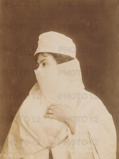 Portrait of an unknown veiled Turkish woman, Dame Turque voilée (profil), photograph, number: 'No 112', photographer: G. Lekegian & Co., Middle East, 1887 - c. 1900, paper, albumen print, height 275 mm x width 213 mm, adult woman, anonymous historical person depicted, A sepia-toned portrait of a person turned in profile wearing a light-colored head covering and veil that conceals the lower face, revealing only the eyes and a curl of hair at the forehead; the person’s hand is raised to the chest, showing multiple rings on the fingers and a bracelet at the wrist, and the clothing includes layered fabric with lace trim and a decorative clasp near the waist against a plain, dark background.