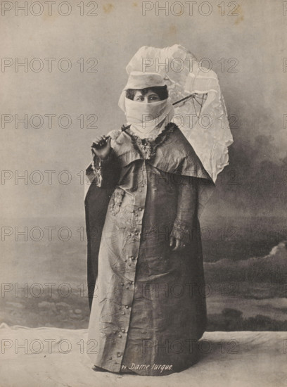 Portrait of an unknown veiled Turkish woman, Turkish lady, photomechanical print, number: '99.', Middle East, cliché maker: anonymous, 1870 - 1930, photographer, paper, collotype, photogravure, height 236 mm x width 180 mm, adult woman, anonymous historical person portrayed, A full-length sepia-toned photograph of a person standing and facing the camera, wearing a long, buttoned outer garment with a shiny, textured surface and a contrasting inner layer; the upper garment has a decorative collar and ruffled trim, and one sleeve shows a cuff. The person's face is mostly covered by a light-colored cloth wrapped around the lower face and head, leaving only the eyes visible under a small brimmed hat. The person holds an open lace-patterned parasol over a shoulder with one hand, while the other hand is raised near chest level holding a small object. The background is a plain, softly shaded backdrop and the floor beneath the figure shows a draped cloth.