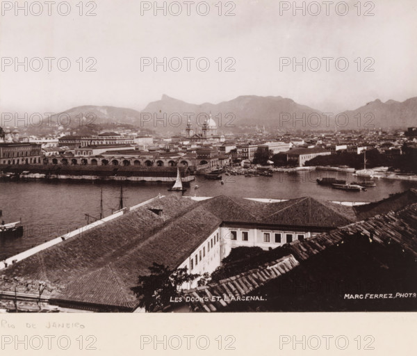 View of the port and the arsenal of Rio de Janeiro, Les docks et l'arsenal, photomechanical print, address: 'MARC FERREZ, RIO DE JANEIRO, 88, RUA SÃO JOSÉ', photographer: Marc Ferrez, Rio de Janeiro (city), cliché maker: anonymous, 1890 - 1910, Marc Ferrez, photographer, cliché maker, Sudeste, pasteboard, paper, collotype, photoprint, height 150 mm x width 202 mm, height 259 mm x width 332 mm, harbour, ammunition store, arsenal, A sepia-toned aerial view of a coastal city showing a waterfront with several boats and sailboats moored in calm water, a long dockside building with arched openings along the water, and a cluster of low-rise buildings with tiled roofs in the foreground. Beyond the waterfront are more multi-story buildings, a prominent church with domes and towers rising above the surrounding structures, and additional architectural details across the harbor. In the background a range of mountains forms a silhouette beneath a pale sky.