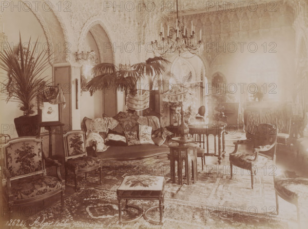 Alger (salon mauresque, palais du 1e President), Interior of the El Mouradia in Algiers, photograph, number: '(2624).', Algiers, 1860 - c. 1900, photographer, North Africa, paper, albumen print, height 188 mm x width 250 mm, palace - AA - civic architecture: inside, El Mouradia, A warmly toned room filled with ornate furnishings and decorative details, showing upholstered chairs with patterned fabric, a cushioned settee draped with various pillows and textiles, small wooden tables with carved legs, and a low cushioned footstool on a richly patterned rug; tall potted palms and other plants are placed around the space, and a multi-armed chandelier hangs from a highly ornamented ceiling with carved arches and intricate wall plasterwork; decorative lamps with fringed shades, framed mirrors, screens, and carved wooden cabinets appear along the walls, and light filters through a large window, creating a soft bright area on one side of the room.