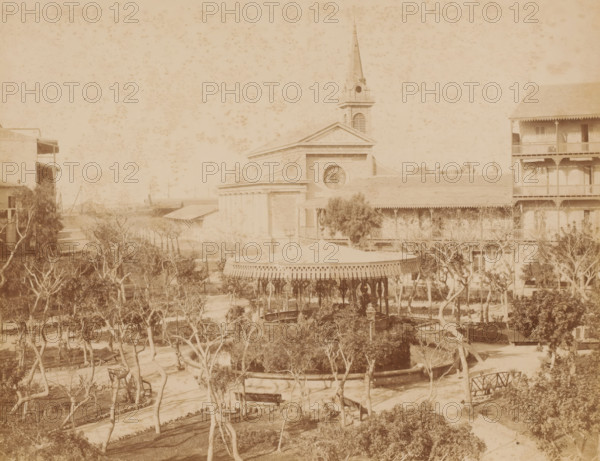 View of the Place de Lesseps in Port Said, Place de Lesseps, Port Saïd, photograph, inscription: 'Place de Lesseps Port Said, photo Zangaki, Port Said', photographer: Zangaki, C. & G., Port Said, 1870 - 1910, North Africa, pasteboard, paper, albumen print, height 219 mm x width 276 mm, height 347 mm x width 453 mm, square, circus, church (exterior), merry-go-round, roundabout, chair o'plane, A faded sepia photograph of a park scene with a central gazebo featuring a decorative roof and ornate pillars surrounded by trees and winding paths, benches placed near the paths, and a circular low wall around a planted area; behind the gazebo is a large classical building with a triangular pediment, a circular window, and a tall steepled tower, flanked on the right by a multi-story building with balconies and on the left by another building with exposed verandas, all seen under a speckled sky.