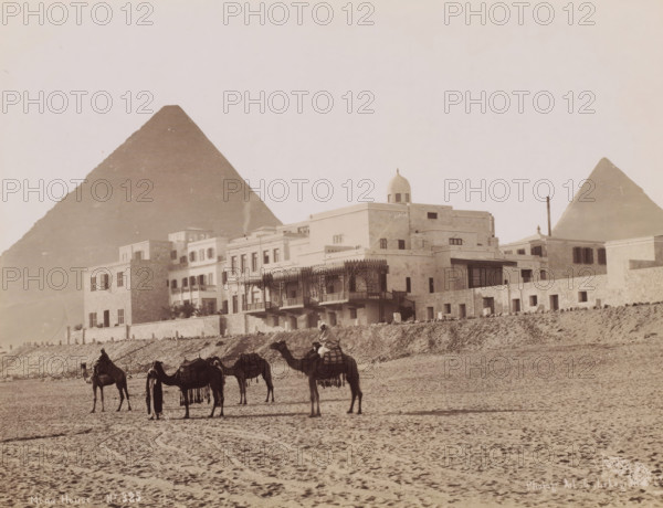 Mena House, View of the Mena House Hotel near Cairo, photograph, number: 'No 325', photographer: G. Lekegian & Co., Cairo (city), 1887 - 1900, Ottoman Empire, North Africa, paper, albumen print, height 211 mm x width 275 mm, hotel, hostelry, inn, pyramid (historical grave form), Mena House Hotel, A wide sandy foreground with visible hoof and foot tracks leads to a row of camels standing and being led by figures, some mounted and some standing beside the animals, the camels carrying saddles and decorative fringes; behind them a low retaining wall separates the sand from a terrace of multi-story stone and stucco buildings featuring balconies, shuttered windows, an ornate wooden veranda, arched openings, and a domed rooftop structure; in the background two large pyramidal stone structures rise against a pale sky.