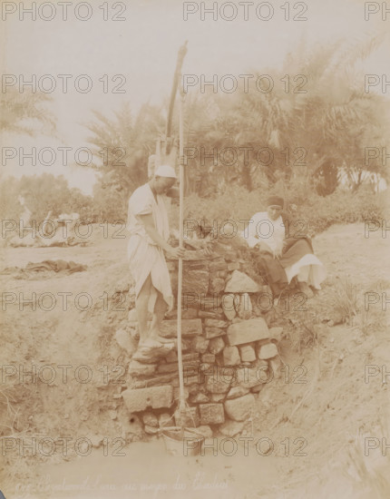 Two unknown men at a water pump, Elévation de l'eau au moyen du Chadou, photograph, number: '646', photographer: Félix Bonfils, Middle East, 1867 - 1885, Félix Bonfils, photographer, paper, albumen print, height 282 mm x width 222 mm, utilitarian, public pump or water fountain, A sepia-toned scene shows a man standing on a rough stone structure holding a long vertical pole that extends down into a bucket at the base, another man sits on the stonework nearby wrapped in layered garments and a cloak, palm trees and dense foliage fill the background behind them, the foreground includes uneven dirt ground and scattered stones, and a faint body of water is visible at the bottom where the bucket rests.