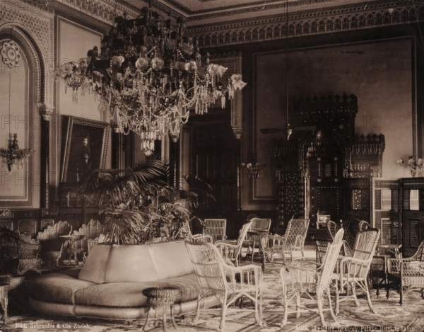 Le Caire, Gezireh, Palace Hotel, le Salon, Salon in the Gezirah Palace Hotel in Cairo, photomechanical print, number: '5125.', Gezirah, clichémaker: anonymous, publisher: Schröder & Co., Zurich (city), 1894 - 1895, photographer, Ottoman Empire, North Africa, Cairo (city), paper, collotype, photogravure, height 211 mm x width 269 mm, hotel, hostelry, inn, drawing-room, 'salon', Gezirah Palace, A large ornate chandelier with hanging crystals dominates the ceiling area above an elaborately decorated room with patterned cornices and framed wall panels; beneath it are wicker chairs and small tables arranged around a low upholstered circular sofa and a round woven stool, with potted palms and other plants placed near the seating; carved wooden screens and an intricately carved niche with geometric and arched details occupy the back wall, while framed artwork leans against a side wall and wall-mounted light fixtures with glass shades flank the room.
