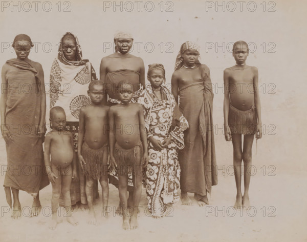 Femmes et enfants des regions equitoriales, Group portrait of nine unknown women and children from French Equatorial Africa, photograph, number: 'N 307', photographer: G. Lekegian & Co., Africa, 1887 - 1900, paper, albumen print, height 212 mm x width 276 mm, anonymous historical persons depicted in a group, in a group-portrait, French Equatorial Africa, A group of people standing in a row against a plain light background, wearing a variety of cloths and wraps; some are clothed in patterned fabric draped around their bodies, others in plain wraps, and two children wear fringed skirts; a few wear headwraps or scarves, and one has a patterned shawl covering much of the body; skin tones appear dark and hair styles vary from short to braided; most stand barefoot on a light ground and face the camera with neutral expressions.