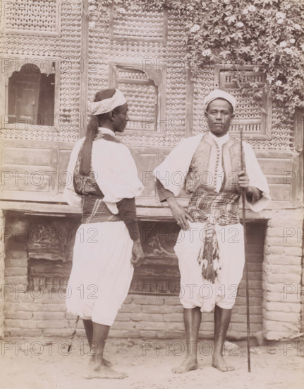 Double portrait of two unknown Egyptian soldiers (Séis), Séis, photograph, number: '289 ?', photographer: Jean Pascal Sébah, Egypt, 1888 - 1900, North Africa, paper, albumen print, height 270 mm x width 212 mm, the soldier, the soldier's life, Two men stand barefoot in front of a patterned wooden wall with carved panels and a flowering vine at the top right; one man faces slightly away showing a wrapped headscarf, long hair tied at the back, a patterned vest over a light shirt, and a cloth sash around his waist, while the other faces forward holding a long staff, wearing a wrapped head covering, an embroidered vest over a light shirt, a patterned sash tied at his waist with tassels, and loose light trousers.