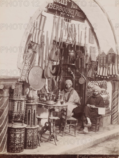 Barbouchi Frères in front of their shop in the Souk el-Trouk in the Medina of Tunis, Boutique de Barbouchi, photograph, number: 'N. 70', photographer: J. André Garrigues, Medina of Tunis, 1860 - 1900, North Africa, Tunis, paper, albumen print, height 258 mm x width 199 mm, shop, store, Souk el-Trouk, A storefront framed by a rounded arch displays hanging rifles and long wooden items, decorative shields, patterned textiles, and ornate metal lanterns and vessels; two men wearing wrapped head coverings and layered clothing sit among stacked folded fabrics and carved wooden tables and stools with inlaid designs, surrounded by small metal pitchers, trays, and intricately patterned boxes and panels.