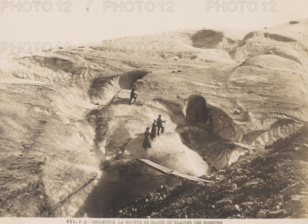 Chamonix, la Grotte de Glace du Glacier des Bossons, Hikers on the Bossons Glacier in Chamonix, photograph, number: '651.', publisher: Photochrom Zürich, Zürich (city), 1889 - c. 1920, photographer, paper, baryta paper, gelatin silver print, developing gelatin silver print, height 165 mm x width 227 mm, walking, hiking (recreation), glacier, Bossons Glacier, A pale, textured icy landscape with undulating snow and deep crevices is shown, featuring a group of people wearing long coats and hats standing and walking on the ice, some holding walking sticks, one person positioned higher on a ridge and another silhouetted inside a dark ice cave opening, a narrow wooden plank bridge spans a crevasse near rocky ground at the lower edge, and scattered rocks and shadowed hollows punctuate the icy surface under a bright, empty sky.