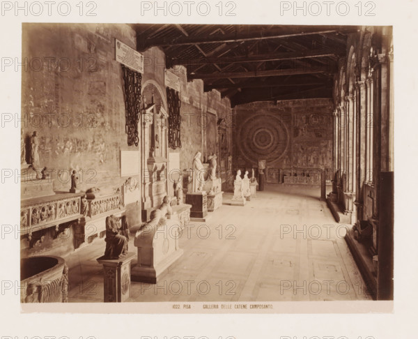 Sculpture gallery in the Camposanto in Pisa, Italy, Galleria delle Catene Camposanto, Pisa, photograph, number: '1022.', after sculpture by various manufacturers, 1851 - 1900, various manufacturers, photographer, after sculpture by, Tuscany, various makers, pasteboard, paper, albumen print, height 310 mm x width 383 mm, gallery of sculptures, Campo Santo (Pisa), A long colonnaded interior space with a high wooden beamed ceiling and a row of arched openings along the right side, stone floor tiles, and walls decorated with large circular and figurative wall paintings. Along the left wall there are sculpted tombs, ornate carved panels, and freestanding statues and busts on pedestals, with decorative reliefs and sculpted figures visible. Two large heavy chains hang beside a recessed architectural niche with an ornate frame and sculpted elements. Several sculpted reclining figures and seated figures rest atop tomb structures, and a freestanding column-like object stands near the far wall be...