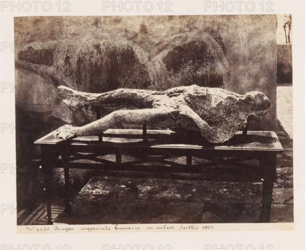 Empreinte humaine un esclave fouilles 1863, Plaster cast of a body in Pompeii, Italy, Pompei, photograph, number: '21', number: '11', number: '16', number: 'No. 4235', photographer: attributed to Giorgio Sommer, Pompeii, 1863 - 1875, Attributed to, pasteboard, paper, albumen print, height 304 mm x width 403 mm, man in a general biological sense, A horizontal image showing a pale, textured human-shaped form lying face up on a rectangular table with visible legs and cross supports, the form appearing rough and mottled against a darker, uneven background with arch-like markings and varied surface textures, the table set on a stone or tiled floor with a patterned area partially visible, and a narrow vertical strip at the right edge revealing a lighter outdoor area with a small tree silhouette.