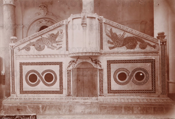 Ravello - Pta. Giona, Pulpit in the cathedral of Ravello, photograph, number: 'N. 49', Ravello, 1880 - 1920, photographer, paper, daylight gelatin silver print, height 141 mm x width 260 mm, pulpit, ambo, chancel, Dom (Ravello), A decorative stone or plaster façade with symmetrical rectangular panels and patterned borders fills the image, featuring two stylized winged creatures facing inward along the upper sloped panels and a small human-like figure near one creature, a central vertical column with a carved eagle at its top, arched recessed doorway beneath the column flanked by bird motifs, circular window-like openings set inside figure-eight patterned designs on each lower panel, fluted columns visible at the far left and right edges, and carved ornamental molding and a patterned base along the bottom.