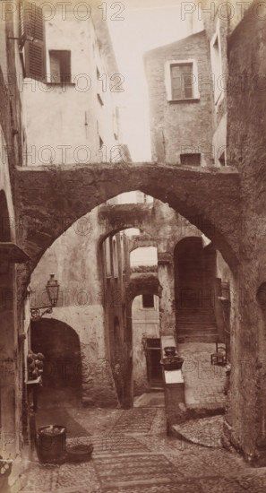 Alley in San Remo, San Remo, photograph, 1851 - 1900, photographer, paper, albumen print, albumendruk, height 271 mm x width 148 mm, street, A narrow, cobbled alleyway framed by tall, weathered stone and plaster buildings with multiple arched passageways and stone bridges crossing overhead; the walls show textured, worn surfaces and small windows with shutters and simple frames, one shutter partially open; a wrought iron street lamp projects from a wall near an arch; stone steps and uneven paving lead down the alley, where clay pots, basins, and a metal bucket sit on ledges and the ground; a simple wooden chair rests against a wall near a doorway and a small open arch reveals deeper recesses and stairways within the buildings.