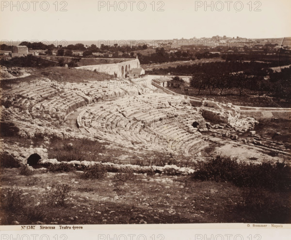 Teatro greco, View of the ruins of the Greek amphitheater in Syracuse, Sicily, Siracusa, photograph, number: '22', number: 'No. 1382', photographer: Giorgio Sommer, Syracuse (city), 1857 - 1914, pasteboard, paper, albumen print, height 310 mm x width 384 mm, ruin of a building, architecture, amphitheatre, A wide sepia photograph showing the stone remains of a large semicircular amphitheater with concentric carved seating and worn pathways, surrounded by rough grassy terrain and scattered rocks. Behind the amphitheater a low stone wall and an arched aqueduct or bridge lead toward small buildings and a rural landscape of trees and fields. In the distant background a town with clustered buildings and a skyline of varied rooflines sits along the horizon beneath a pale sky.