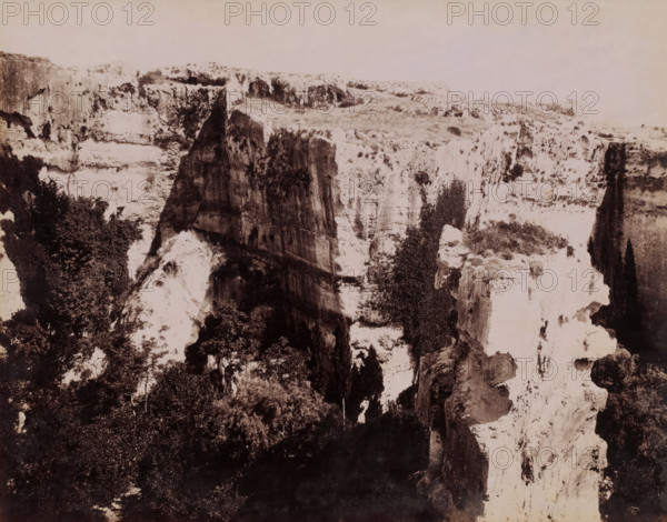 View of Latomie del Paradiso in Syracuse, Sicily, Latomie del Paradiso, Siracusa, photograph, number: '2652', photographer: Giorgio Sommer, Syracuse (city), 1857 - 1914, pasteboard, paper, albumen print, height 198 mm x width 250 mm, quarry, A sepia-toned photograph of a deep rocky canyon with steep vertical cliffs and jagged rock faces, textured surfaces showing striations and erosion, patches of vegetation clinging to ledges and crevices, darker shadowed recesses between the cliffs, a plateau of lighter rock and sparse scrub at the top, and dense clusters of trees and bushes at the canyon base.