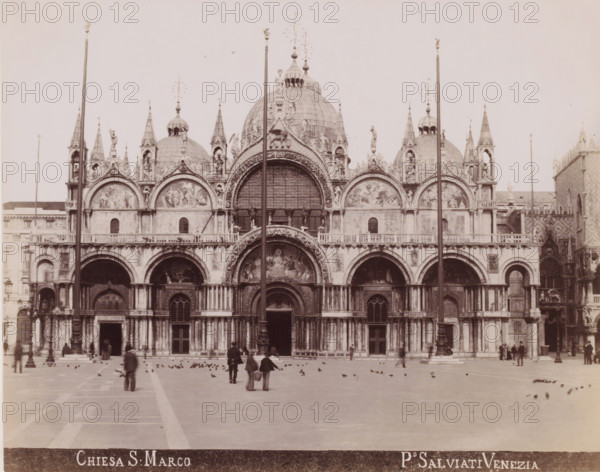 View of San Marco in Venice, Chiesa S. Marco, photograph, inscription: 'Venetie', photographer: Paolo Salviati, Venice, 1860 - 1870, pasteboard, paper, albumen print, albumendruk, height 199 mm x width 251 mm, height 275 mm x width 365 mm, square, circus, church (exterior), St. Mark's Basilica, St. Mark's Square, A wide facade of an ornately decorated cathedral fills the image, featuring multiple domes, pointed spires, arched doorways, and sculpted reliefs above the arches; a large central arch with detailed artwork sits above the main entrance, flanked by smaller arches and rows of columns; tall flagpoles rise in front of the building; a broad open plaza extends from the cathedral with scattered birds on the ground and several people walking or standing, some in groups and some alone; the overall image is monochrome with high contrast and an expansive sky above the building.