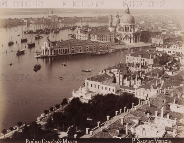 Pan:a dal Camp:e di S: Marco, View of Venice from the Campanile of San Marco, photograph, inscription: 'Venetie', photographer: Paolo Salviati, Venice, 1860 - 1870, pasteboard, paper, albumen print, albumendruk, height 196 mm x width 250 mm, height 276 mm x width 365 mm, city-view in general, 'veduta', canals, waters (in city), Grand Canal, Basilica of San Marco, A wide aerial view of a waterfront cityscape showing a large domed church with ornate facades near a quay lined with a long stone building with arched openings, many adjacent multi-story buildings with tiled roofs and chimneys, and a waterfront area populated by numerous moored sailboats and small rowboats on calm water, with a tree-lined promenade and garden area visible in the foreground and a distant shoreline and low buildings across the water.