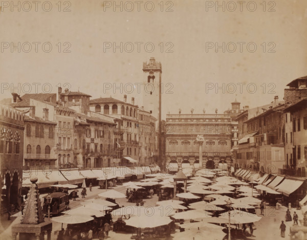 Market square in Verona, Italy, photograph, number: '13', inscription: 'Verona', inscription: 'Verona - Mercato ... Erbe ?', Verona, 1851 - 1900, photographer, pasteboard, paper, albumen print, albumendruk, height 304 mm x width 404 mm, market, square, circus, A crowded open market fills the foreground with rows of shaded stalls topped by large umbrellas and canopies, and people walking or standing among them; on the left a stone obelisk with carved details rises from a pedestal next to a covered tram and a small group of figures; surrounding the market are multi-story buildings with tiled roofs, balconies, arched windows, and various chimneys, and a tall square tower with an open belfry stands near the center background adjacent to an ornate façade decorated with sculpted figures and reliefs; the sky above is pale and largely empty.