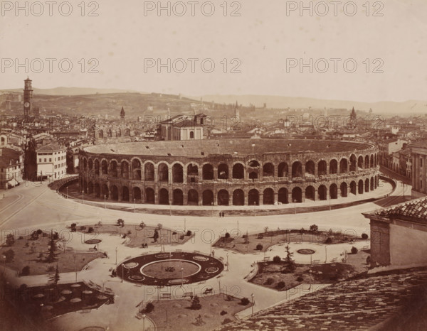 View of the Arena of Verona, Italy, photograph, inscription: 'Verona.', number: '13', Verona, 1851 - 1900, photographer, pasteboard, paper, albumen print, height 306 mm x width 405 mm, city-view in general, 'veduta', amphitheater, Arena of Verona, A wide sepia-toned cityscape centered on a large oval stone amphitheater with multiple tiers of arched openings and a curved exterior roadway, surrounded by low buildings and narrow streets; in front of the amphitheater is a landscaped plaza with circular flowerbeds, paved paths, benches, streetlamps, and small trees, with a few scattered people walking or sitting; beyond the amphitheater a dense cluster of historic buildings and towers stretches toward rolling hills in the background, and the sky above is pale and largely empty.