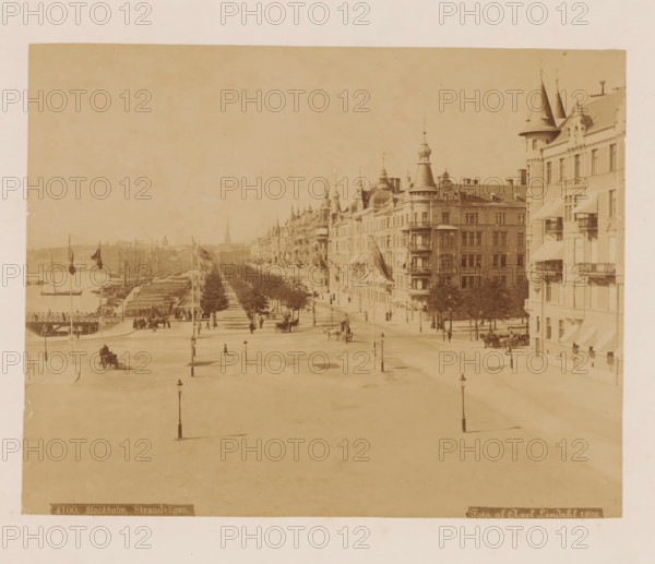Stockholm, strandvagen, View of Strandvägen and the harbor of Stockholm, photograph, number: '4100.', photographer: Axel Lindahl, 1898, pasteboard, paper, albumen print, albumendruk, height 166 mm x width 213 mm, height 345 mm x width 455 mm, harbour, avenue, boulevard, promenade, esplanade, Strandvägen, A wide sepia-toned street scene viewed from above showing a broad open square foreground with tall street lamps and sparse pedestrians, a tree-lined promenade extending toward the background flanked by ornate multi-story buildings with turrets, balconies, and awnings on the right, flags flying from poles along the promenade, horse-drawn carriages and people along the roadway, a waterfront with docked sailboats visible to the left, and a distant skyline with spires and low buildings under a pale sky.