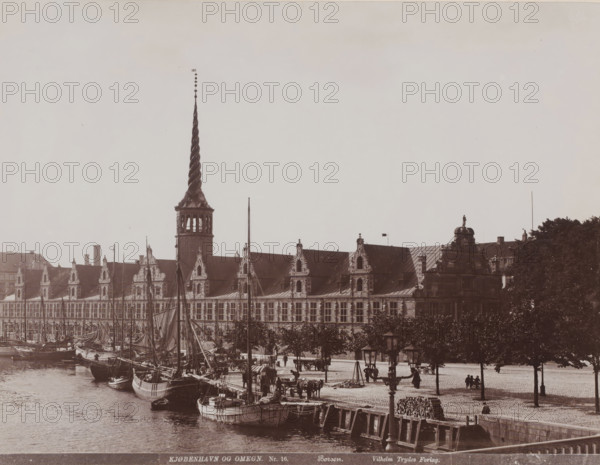 Børsen on Slotsholmen in Copenhagen, left of center the Dragespiret, Børsen, Kjøbenhavn og Omegn, photograph, number: 'Nr. 16.', Copenhagen (city), publisher: Vilhelm Trydes, Denmark, 1880 - 1910, photographer, paper, pasteboard, baryta paper, daylight collodion silver print, height 351 mm x width 457 mm, stock exchange, stock-market, Slotsholmen, A waterfront scene showing a long ornate building with a roofline punctuated by decorative gables and a tall twisted spire rising from a tower, boats moored along a quayside with masts and rigging, a cobblestone promenade with lamp posts and stacked logs, several people and horse-drawn carts near the water, and trees lining the promenade on the right.