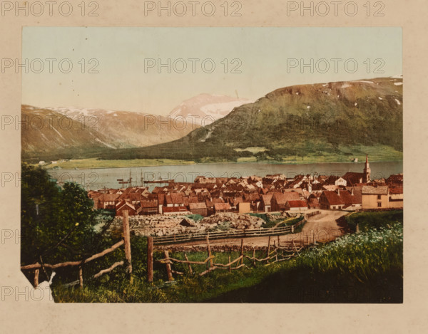 Village view in Norway, photomechanical print, Norway, cliché maker: anonymous, 1860 - 1900, photographer, pasteboard, paper, albumen print, color lithography, albumen printing with color lithography, height 165 mm x width 228 mm, height 349 mm x width 457 mm, city-view in general, 'veduta', A panoramic view shows a village with clustered houses topped by reddish-brown roofs and light-colored walls, a church with a tall steeple near the water, a shoreline with boats and masts visible on the calm water, rolling green fields and fenced pastures in the foreground with a dirt path leading toward the village, dense trees and shrubs near the front left, and distant mountains with patches of snow under a pale sky.
