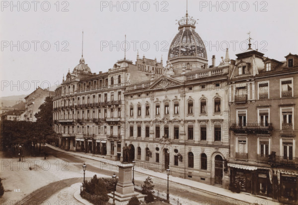 Street view in German city (former title), View of Leopoldsplatz in Baden-Baden, photograph, In the middle the post office ('Kaiserliches Postamt') and next to it Hotel Holland., check stamp: '1903, Gust. Salzer, B. Baden', number: '125', photographer: Gustav Salzer, Baden-Baden, 1903, Baden-Württemberg, paper, baryta paper, daylight collodion silver print, height 115 mm x width 164 mm, square, circus, hotel, hostelry, inn, post-office, A wide street scene shows a row of ornate multi-story stone and brick buildings with decorative facades, balconies with wrought iron railings, and numerous tall windows with curtains; a large domed tower with an intricate lattice structure rises above one building, flanked by smaller domes and spires topped with finials; sculptural figures and ornamental stonework decorate the rooflines and pediments; a broad sidewalk runs along the buildings and a tree-lined area with mature trees and shrubs sits opposite; a stone monument with a statue atop a tall ped...