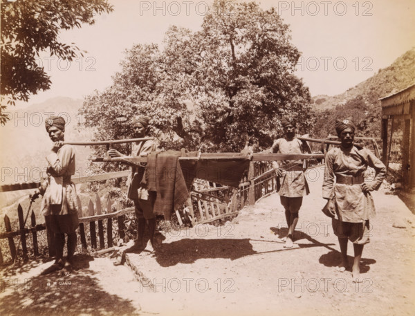 Four Indian men in unform life a palanquin, India, photograph, signature: 'SH Dagg', inscription: '£1', inscription: 'Asia', photographer: Sidney Herbert Dagg, British India, 1890 - 1910, pasteboard, paper, albumen print, height 212 mm x width 273 mm, height 227 mm x width 292 mm, (carrying a person on a) litter, stretcher, Four men wearing turbans and tunics stand on a dirt path beside a wooden fence and railing, two of them holding a long wooden pole across their shoulders that supports a stretcher-like frame draped with a cloth, while a large tree with dense foliage fills the background and a hillside and a simple building with a slanted roof appear to the right.