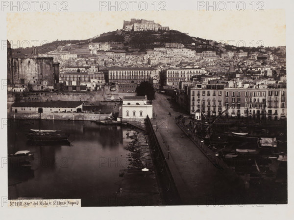 View of the Sant'Elmo castle on Mount Vomero in Naples, Italy, Strada del Molo v. St. Elmo, Napoli, photograph, number: 'No. 1112.', number: '22', inscription: 'Naples.', address: 'GIORGIO SOMMER, STUDIO MONTE DI DIO 4., MAGAZZINO S. CATERINA 5., NAPOLI', photographer: Giorgio Sommer, Naples, 1890 - 1910, Giorgio Sommer, photographer, pasteboard, paper, albumen print, height 307 mm x width 377 mm, city-view in general, 'veduta', castle, Castel Sant'Elmo, A black and white panoramic view of a waterfront and an urban hillside showing a harbor with moored boats and masts, a long quay lined with lamp posts and a paved road, waterfront buildings with multiple stories and balconies, a lighter colored rectangular building close to the water, a large round stone fortification on the left, dense rows of varied buildings climbing the hillside, terraces and trees on the slope, and a prominent castle-like structure perched atop the hill.