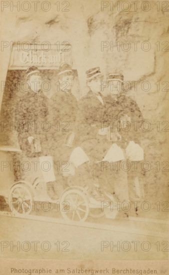 Group portrait of four women wearing caps, sitting on a carriage on a rail, cabinet photograph, name: 'P. Ney Atelier für Photographie am Königl. Bayerischen Salzbergwerk Berchtesgaden, ', photographer: Paul Ney, Berchtesgaden, c. 1870 - c. 1900, Bavaria, paper, pasteboard, albumen print, height 148 mm x width 97 mm, adult woman, railway, train, A faded sepia photograph showing four people seated side by side on a small wooden cart with visible spoked wheels, each person wearing a dark jacket and a cap with a light band, lighter cloth draped across their laps, and small hand lanterns held or attached near their hands, with a rectangular signboard partially visible behind them and a mottled background surrounding the group.