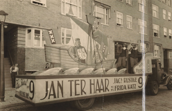 Decorated float of the political party List 9 during a parade in Amsterdam, photograph, postcard, annotation: 'Amsterdam, 1930s', photographer: T en M, Amsterdam, 1920 - 1940, paper, gelatin silver print, height 89 mm x width 139 mm, political parties, public festivities (parade, pageant, cavalcade, festive activities), A street scene showing a large parade float on a flatbed truck in front of a brick apartment building, the float featuring a painted wooden ship with a tall sail, a painted figure on the ship holding a shield, decorative stylized waves along the float base, two costumed men standing on the float blowing horns, a man leaning against a doorway at the building entrance and another person partially visible near a window behind the float, multi-pane windows with curtains across the building facade, a lamppost at the left edge, and cobblestone pavement beneath the truck.