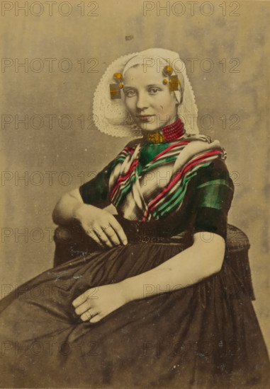 Portrait of an unknown woman in traditional costume of Goes, Zeeland, cabinet photograph, annotation: 'Goes, R..., Costumes des Pays Bas., Publié par A. Jager, Damrak 81, Amsterdam', annotation: 'Zeeland traditional costume.', number: '40', photographer: Andries Jager (possibly), Amsterdam, 1860 - 1890, paper, pasteboard, albumen print, hand coloring, height 156 mm x width 115 mm, folk costume, regional costume, historical persons not known by name - BB - woman, A seated young woman wearing a lace headpiece with rounded edges and decorative adornments at the temples, a striped shawl with bright narrow bands draped over a dark short-sleeved bodice, and a high necklace of small beads; she has a composed facial expression and is looking toward the viewer, one arm resting on an armrest and the other hand placed on her skirt, with rings visible on fingers and a pleated dark skirt extending across her lap.