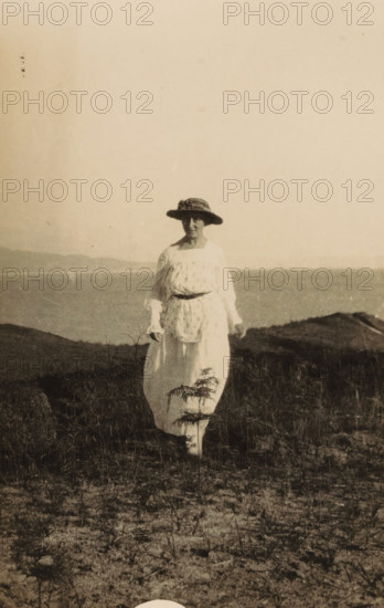 Portrait of an unknown woman near a coast, photograph, inscription: 'Voor Kees, from aunt Cor', 1919-09-28 - 1929-09-28, photographer, paper, baryta paper, gelatin silver print, developing gelatin silver print, height 135 mm x width 86 mm, anonymous historical persons in a double portrait - BB - woman, A sepia-toned photograph of a person standing outdoors on grassy, sandy ground near a body of water, wearing a light-colored ankle-length dress with a patterned apron or overdress and a wide-brimmed hat with a decorative band, the person facing the camera with hands relaxed at their sides, low hills visible across the water in the background and sparse vegetation in the foreground.