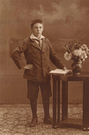 Studio portrait of an unknown boy next to a table, photograph, postcard, photographer: Machiel Hendricus Laddé, Amsterdam, c. 1907 - c. 1920, paper, gelatin silver print, developed gelatin silver print, height 133 mm x width 89 mm, boy (child between toddler and youth), cut flowers, nosegay, bunch of flowers, table, A sepia-toned portrait of a young person standing with one hand on a hip and the other resting on an open book placed on a round wooden table, wearing a dark jacket with a wide white collar and a patterned tie, short trousers, long dark socks, and leather shoes; a decorative vase of mixed flowers sits on the table beside the book, and a patterned rug covers the floor beneath them against a softly blurred backdrop.