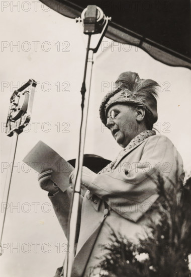 HM the Queen opens the World Jamboree July 31, 1937, photograph, postcard, Vogelenzang (North Holland), publisher: Have, W. ten, Amsterdam, 1937-07-31, photographer, paper, gelatin silver print, height 151 mm x width 105 mm, Person, Wilhelmina of the Netherlands, queen, A black-and-white image of an older person wearing round glasses and a feathered hat, shown from a low angle behind a microphone stand, holding a sheet of paper and dressed in a coat with a patterned collar.