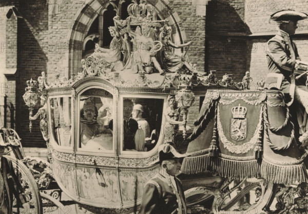 On the way to the Groote Kerk, for the baptism of Princess Beatrix, photograph, postcard, publisher: Have, W. ten, Amsterdam, 1938-05-12, photographer, printer (person), paper, gelatin silver print, height 105 mm x width 149 mm, Person, Bernhard (Prince of the Netherlands), Beatrix (Queen of the Netherlands), Juliana of the Netherlands, prince, princess, crown princess, A richly decorated carriage with ornate sculpted figures on its roof and carved details along its sides is shown in front of a brick building with arched windows; the carriage windows reveal people inside wearing formal headwear and garments, and a uniformed rider sits at the front holding reins while another uniformed attendant stands nearby; heavy draped cloths with embroidered motifs and tasseled trim hang over the carriage, and detailed metal and sculptural ornamentation, including crowns and human-form figures, adorn the vehicle.