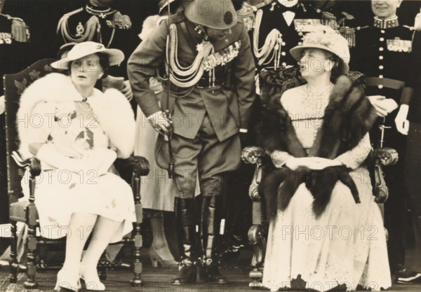 Army Day in The Hague, photograph, picture postcard, The Hague, publisher: Have, W. ten, Amsterdam, 1938-08-29, photographer, printer (person), paper, gelatin silver print, developing gelatin silver print, height 105 mm x width 150 mm, Person, Juliana of the Netherlands, Wilhelmina of the Netherlands, queen, crown princess, A black-and-white photograph showing two seated women in ornate chairs and a uniformed man standing between them; the woman on the left wears a light-colored dress with a fur stole, a wide-brimmed hat, and white gloves, her legs crossed and shoes visible, while the woman on the right wears a patterned dress with a fur wrap, a decorated hat, and gloves, her hands clasped in her lap; the man between them wears a military-style uniform with decorative cords across his chest, a helmet, high boots, and holds a sword; behind them are other uniformed figures with visible epaulettes and medals.