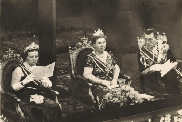 A few days before the 40th anniversary of the reign of HM the Queen. Evening of Honour in the Building for Arts and Sciences in The Hague, photograph, postcard, The Hague, publisher: Have, W. ten, Amsterdam, 1938-08-31, photographer, printer (person), paper, gelatin silver print, height 105 mm x width 150 mm, Person, Bernhard (Prince of the Netherlands), Juliana of the Netherlands, Wilhelmina of the Netherlands, queen, prince, crown princess, Building for Arts and Sciences (The Hague), Three seated people wear formal attire and crowns while sitting on ornate chairs; the left person reads from a folded paper, wearing long gloves and a dark dress with a sash, the central person faces forward with a patterned dress, gloves, a sash, and a necklace, and the right person wears a decorated uniform with braided cords and holds papers. The chairs have carved, decorative backs, and a patterned carpet is visible beneath them. There is a floral arrangement on a low table in front of the central pe...