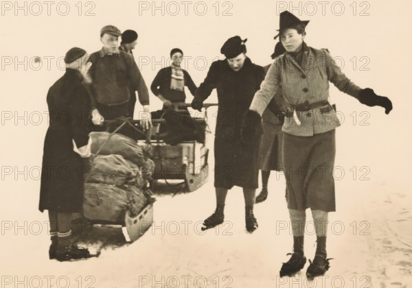 Gouwzee, 23 January 1940, photograph, picture postcard, publisher: Have, W. ten, Amsterdam, 1940, photographer, printer (person), paper, gelatin silver print, developing gelatin silver print, height 104 mm x width 149 mm, skates (winter sports), A group of people dressed in winter clothing stand on snow with sleds and packed bundles; one person in a striped jacket and gloves gestures with an outstretched arm while wearing a pointed hat, another in a dark coat and beret looks downward holding a sled handle, a person with a scarf stands near the center behind the sled, two figures near the left stand beside a loaded sled with bundled items, and the lower halves of skates and winter boots are visible against the snowy ground.