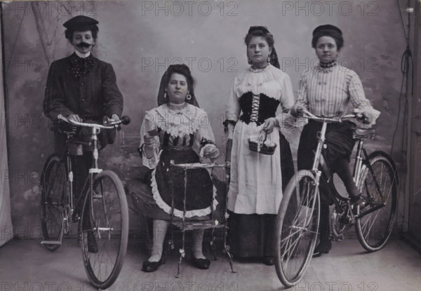 Group portrait of four unknown girls in traditional costume with bicycles, photograph, inscription: 'Studio Arnhem Pastoorstraat 9', photographer: Studio, Arnhem, c. 1895 - c. 1915, paper, pasteboard, height 97 mm x width 140 mm, folk costume, regional costume, bicycle, anonymous historical persons portrayed in a group, in a group-portrait, Four women posed in a studio with two bicycles, two women sitting and two standing, wearing period dresses with lace and high collars, one seated woman holding a small object and wearing striped stockings, one standing woman holding a small woven basket with flowers, two women seated at bicycle handlebars, one woman wearing a dark jacket and hat with a false mustache, hairstyles pinned up, earrings visible on some, and a painted backdrop behind them.