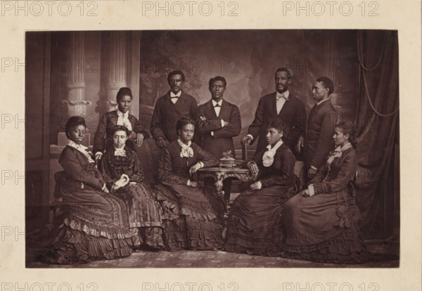 Group portrait of the Jubilee Singers, page, photomechanical print, cliché maker: anonymous, 1872 - in or before 1877, photographer, paper, Woodburytype, height 82 mm x width 130 mm, Print opposite title page., Person, Edmund W. Watkins, Hinton Alexander, Mabel Lewis, Frederick J. Loudin, Thomas Rutling, Maggie Carnes, Maggie Porter, Jennie Jackson, Ella Sheppard, America W. Robinson, historical persons, female singer, male singer, A sepia-toned studio portrait showing a group of adults arranged around an ornate wooden table and upholstered chairs, with decorative columns and drapery in the painted backdrop. The group includes men standing behind and women seated in front, all wearing formal Victorian-era clothing such as dark suits with bow ties for the men and long, layered dresses with ruffles, lace collars, and decorative trims for the women. Several women have their hands resting on the table or on their laps, and one man stands with his arms crossed. Facial expressions are neutra...
