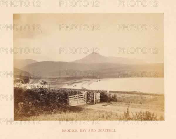 View of Brodick Bay, with Goat Fell in the background, Brodick Bay and Goatfell, photograph, page, photographer: William Russell Sedgfield, Brodick, c. 1861 - in or before 1871, Arran, paper, albumen print, height 81 mm x width 113 mm, gulf, bay, mountain-top, peak, Goat Fell, A faded sepia coastal scene showing a shoreline curving along calm water with small boats near the beach, distant low hills and a prominent conical mountain in the background, foreground vegetation and grassy land leading to a wooden gate and fence at a dirt path, scattered buildings partially obscured by shrubs along the shore, and a wide pale sky above.