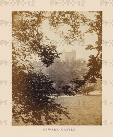 View of Newark Castle, Newark Castle, photograph, page, photographer: William Russell Sedgfield, Selkirk, c. 1862 - in or before 1872, paper, albumen print, height 108 mm x width 85 mm, landscape with tower or castle, A sepia-toned photograph framed by a pale border showing tree branches and foliage in the foreground that form a partial canopy, with open grassy ground below and a distant stone building with battlement-like features perched on a hill, partially obscured by mist or haze and surrounded by more trees.